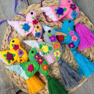 May include: A collection of six felt bird ornaments in pink, blue, yellow, green, and gray, each featuring embroidered floral patterns, a small black eye, and a dangling yarn tassel. Each bird includes a hanging loop for decorative display.