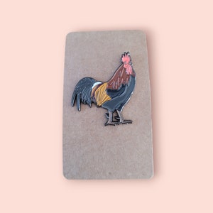May include: A brown cardboard backing with a black and red enamel pin of a rooster. The rooster is facing right and has a red comb and wattle.
