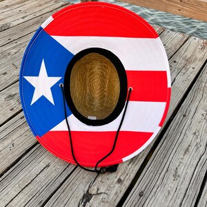 May include: A straw sun hat with a red, white, and blue Puerto Rican flag design. The hat has a black chin strap and is lying on a wooden surface.