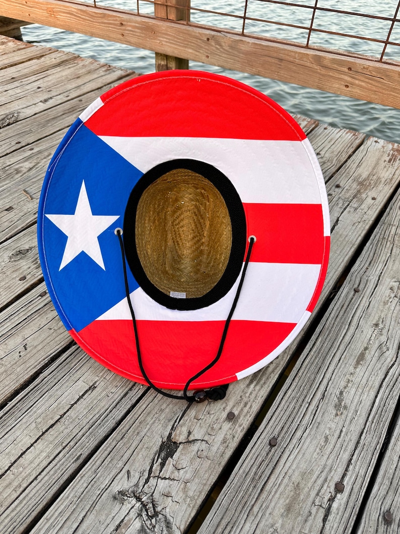 May include: A straw sun hat with a red, white, and blue Puerto Rican flag design. The hat has a black chin strap and is lying on a wooden surface.