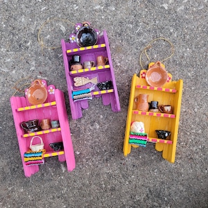 May include: Three miniature wooden wall shelves in pink, purple, and yellow, each featuring three tiers. The shelves are decorated with small painted flowers and hold tiny ceramic pottery, woven baskets, and colorful striped textile pieces.