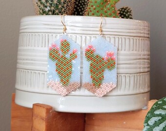 May include: A pair of beaded earrings with a cactus design. The earrings are light blue with green and orange details. The earrings are hanging from a white ceramic planter with a cactus plant inside.