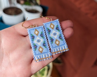 Op de afbeelding: Een paar blauwe kralen oorbellen met een geometrisch patroon in wit, goud en groen. De oorbellen hebben een franje van gouden kralen aan de onderkant.