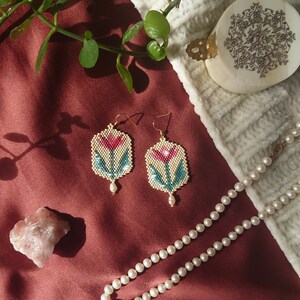 May include: A pair of beaded earrings with a floral design. The earrings are made with white beads and feature a red flower with green leaves. Each earring has a single pearl dangling from the bottom.