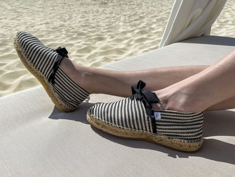 May include: A pair of black and white striped espadrille shoes with black ribbon bows. The shoes are on a white surface.