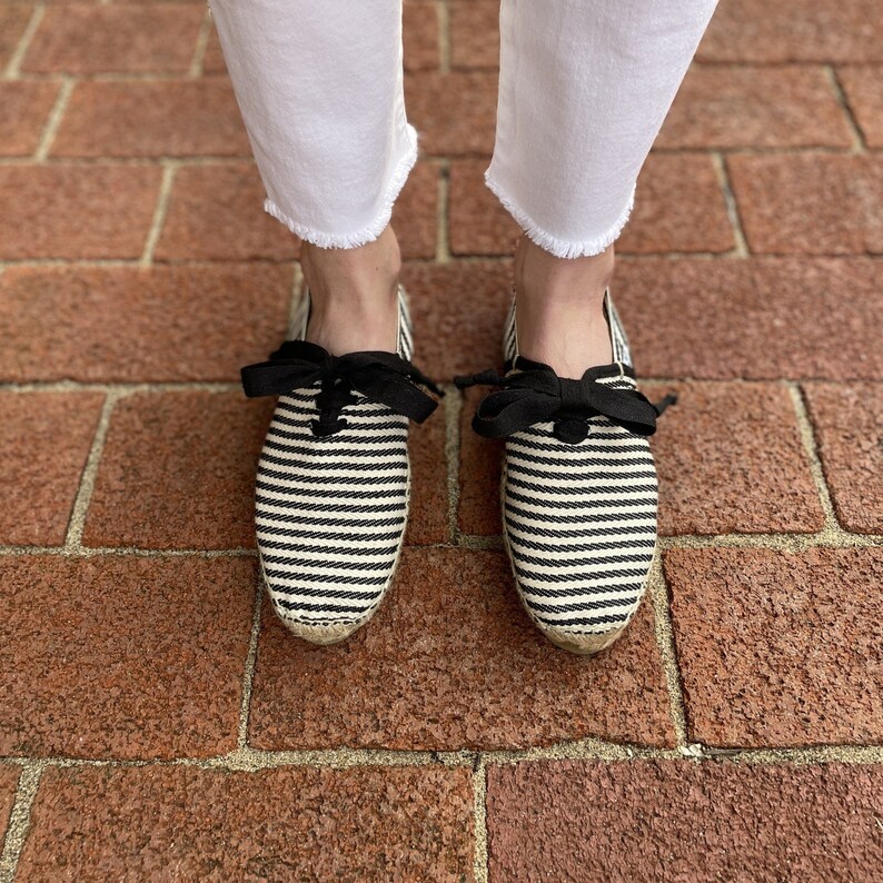 May include: A pair of black and white striped espadrille shoes with black ribbon laces. The shoes are on a brick surface.
