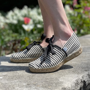 May include: A pair of black and white striped espadrille shoes with black laces. The shoes are made of canvas and have a jute sole.