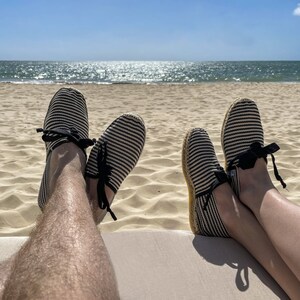 May include: Two people wearing striped espadrille shoes with black ribbon ties, relaxing on a beach with their feet up. The ocean is in the background.