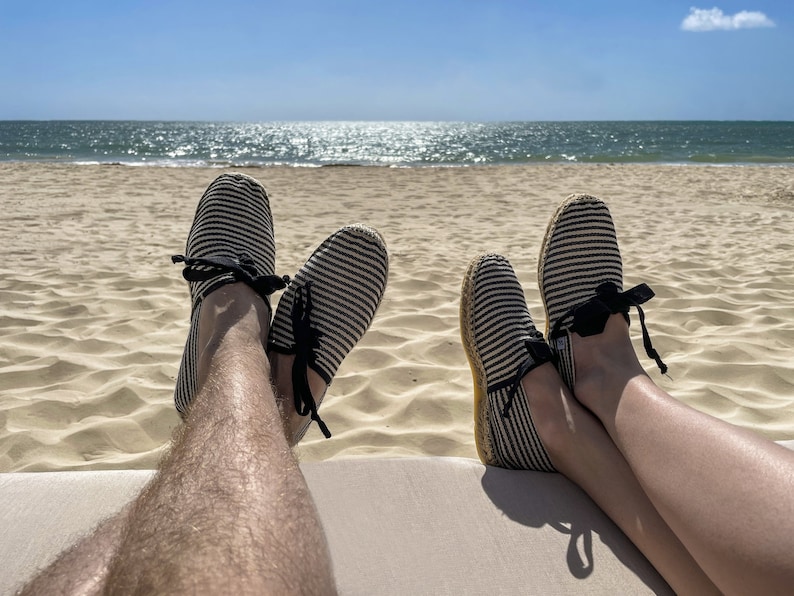 May include: Two people wearing striped espadrille shoes with black ribbon ties, relaxing on a beach with their feet up. The ocean is in the background.