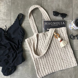 May include: A beige crocheted tote bag with a brown tassel and wooden beads. The bag is lying on a gray tiled floor with a black swimsuit, sunglasses, a magazine, and a tumbler.