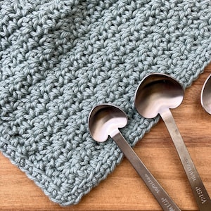May include: A light blue crocheted dishcloth with a textured pattern. Two heart-shaped metal spoons are placed on top of the dishcloth. The spoons have the words "1 tsp" and "1/2 tsp" engraved on them.
