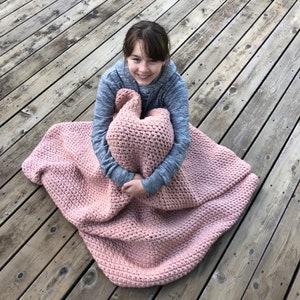 K&ouml;nnte beinhalten: Eine junge Person sitzt auf einem Holzdeck und h&auml;lt eine rosafarbene H&auml;keldecke.