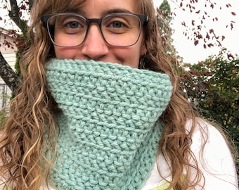 Könnte beinhalten: Ein heller, salbeigrüner, grob gestrickter Schlauchschal mit einer dicken, strukturierten Garnkonstruktion und einem markanten Rippenmuster. Das Accessoire ist so konzipiert, dass es um den Hals getragen wird und eine dichte, warme Schicht bietet.