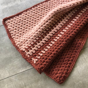 K&ouml;nnte beinhalten: Eine geh&auml;kelte Decke mit einem Farbverlauf von Rosa und Braun. Die Decke ist in der H&auml;lfte gefaltet und die Textur der H&auml;kelstiche ist sichtbar.