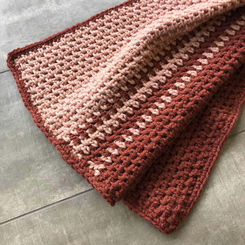 K&ouml;nnte beinhalten: Eine geh&auml;kelte Decke mit einem Farbverlauf von Rosa und Braun. Die Decke ist in der H&auml;lfte gefaltet und die Textur der H&auml;kelstiche ist sichtbar.