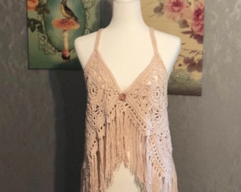 May include: A beige crochet top featuring a V-neckline, thin straps, and a central button closure. The garment is crafted with intricate circular patterns and finished with long, dangling fringe along the bottom hem, displayed on a mannequin.