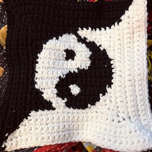 May include: A square, crocheted pillow featuring a black and white yin-yang symbol. The pillow is divided diagonally, with the top left corner in black and the bottom right corner in white. The yin-yang symbol is centered on the pillow.