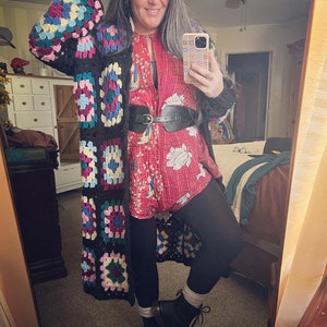 May include: A long, black crochet coat with colorful square patterns is worn over a red floral print dress. The coat features a granny square design with pink, blue, and yellow accents. A black belt cinches the waist, and black leggings and boots complete the look.