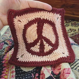 Puede incluir: Un cuadrado tejido a crochet con un diseño de signo de paz rosa y burdeos.