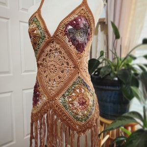 May include: A handmade, tan crochet top with a V-neck and fringe detail. The top features square panels with floral and geometric patterns, incorporating shades of green, burgundy, and tan. The top is displayed on a mannequin.