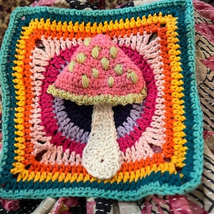 May include: A crocheted square with a colorful rainbow pattern and a white mushroom in the center. The mushroom has a pink cap with green dots and a white stem.