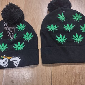 May include: Two black knit beanies with green marijuana leaf patterns. One beanie has a white cartoon hand holding a joint. The beanie has a black pom pom on top and the text "Sports" on the side.