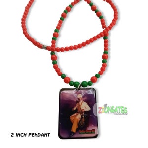 May include: A necklace with a 2-inch pendant featuring an image of a person in a white and gold outfit. The necklace has a red beaded strand with green accents. The pendant has the text "Noble Drew Ali".