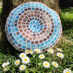Peut inclure: Une pièce circulaire au crochet présentant un motif concentrique de carrés alternés dans des tons bleu clair, turquoise, marron et beige, séparés par une grille blanche. Le design rayonne depuis un point central en un agencement géométrique.