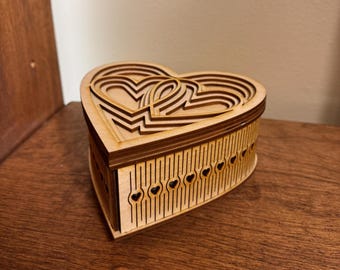May include: A wooden heart-shaped box with intricate laser-cut designs. The box is made of light brown wood and has a layered heart design on the lid. The box is closed and sitting on a wooden surface.