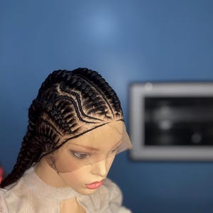 May include: A mannequin head with a black braided wig. The braids are intricately styled, with a combination of straight and curved patterns. The wig is set against a blue background, with a framed picture in the background.