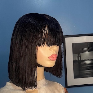May include: A black wig with a blunt cut and fringe is displayed on a mannequin. The wig has a sleek, straight texture and a natural-looking hairline. The mannequin is wearing a light-coloured top with a keyhole neckline. A framed black and white photo hangs in the background.