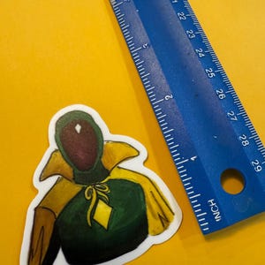 Pu&ograve; includere: Un adesivo verde e giallo di un personaggio che indossa un mantello con cappuccio e un diamante sul petto.