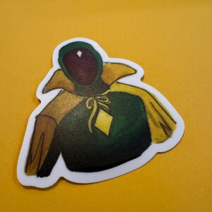 Pu&ograve; includere: Un adesivo con un'illustrazione di un personaggio dei cartoni animati che indossa una tunica verde con cappuccio, un colletto giallo e un diamante giallo sul petto.