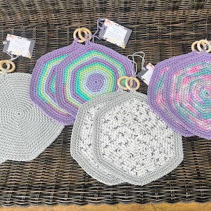 Könnte beinhalten: Eine Sammlung handgefertigter, sechseckiger Topflappen aus Häkelarbeit in verschiedenen Farben. Die Topflappen haben einen Holzring zum Aufhängen und sind in Grau-, Lila- und bunten Mustern erhältlich. Die Topflappen werden auf einer gewebten Oberfläche präsentiert.