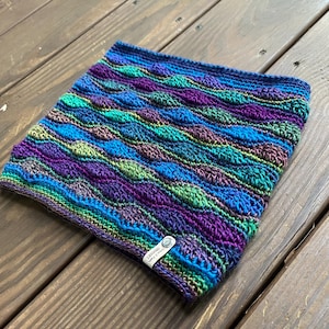 May include: A crochet cowl featuring a textured wave pattern in shades of purple, blue, and green. The accessory includes a small white rectangular tag with the text "LINDSEY CROCHET" attached to the lower edge of the fabric.