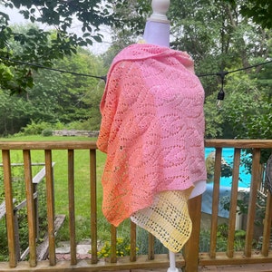 May include: A pink and yellow ombre crochet shawl with a lacy pattern. The shawl is draped over a white mannequin and is displayed on a wooden deck.