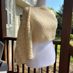 May include: A cream-colored, crocheted bolero with one long sleeve and a cropped design. The intricate lace pattern covers the entire garment, creating a delicate and textured appearance. The bolero is displayed on a white mannequin, showcasing its shape and details.