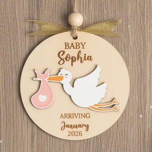 May include: Wooden baby announcement ornament with the name "Sophia", a stork carrying a pink bundle, and the text "Arriving January 2026". The ornament has a gold ribbon and wooden bead for hanging.