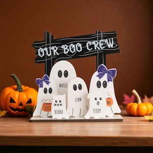 Puede incluir: Decoración de Halloween de madera con un letrero que dice "OUR BOO CREW" encima de cinco figuras de fantasmas blancos. Cada fantasma tiene un nombre y algunos tienen lazos morados. Una calabaza tallada y pequeñas calabazas están en el fondo.