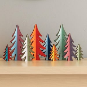 May include: A display of wooden Christmas trees in a range of colours. The trees are painted in shades of red, orange, blue, green, and purple, with white detailing. They are arranged on a light brown surface.