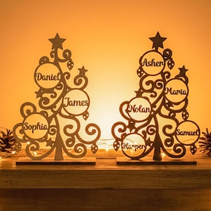 May include: Two wooden Christmas tree decorations with personalised names cut out. The trees have star toppers and decorative cutouts. Names include Daniel, James, Sophia, Asher, Maria, Nolan, Harper, and Samuel. The trees are on wooden bases.