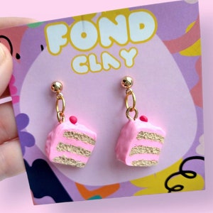 Könnte beinhalten: Rosa und goldene mehrschichtige Kuchenohrringe mit einer roten Kirsche oben drauf. Die Ohrringe befinden sich auf einer Karte mit der Aufschrift "Fond Clay".