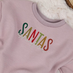 Könnte beinhalten: Mauvefarbenes Sweatshirt mit dem Wort "SANTAS" in bunten Fäden gestickt. Das langärmlige Oberteil hat eine gerippte Textur und einen Rundhalsausschnitt. Das Sweatshirt wird auf einer flauschigen weißen Oberfläche präsentiert.