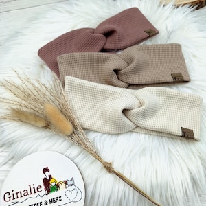 May include: Three waffle-knit headbands in shades of mauve, tan, and cream, each with a twisted knot design. Small brown tags are sewn on the headbands. The headbands are displayed on a white fluffy surface. A wooden sign with text and a drawing is in the foreground.