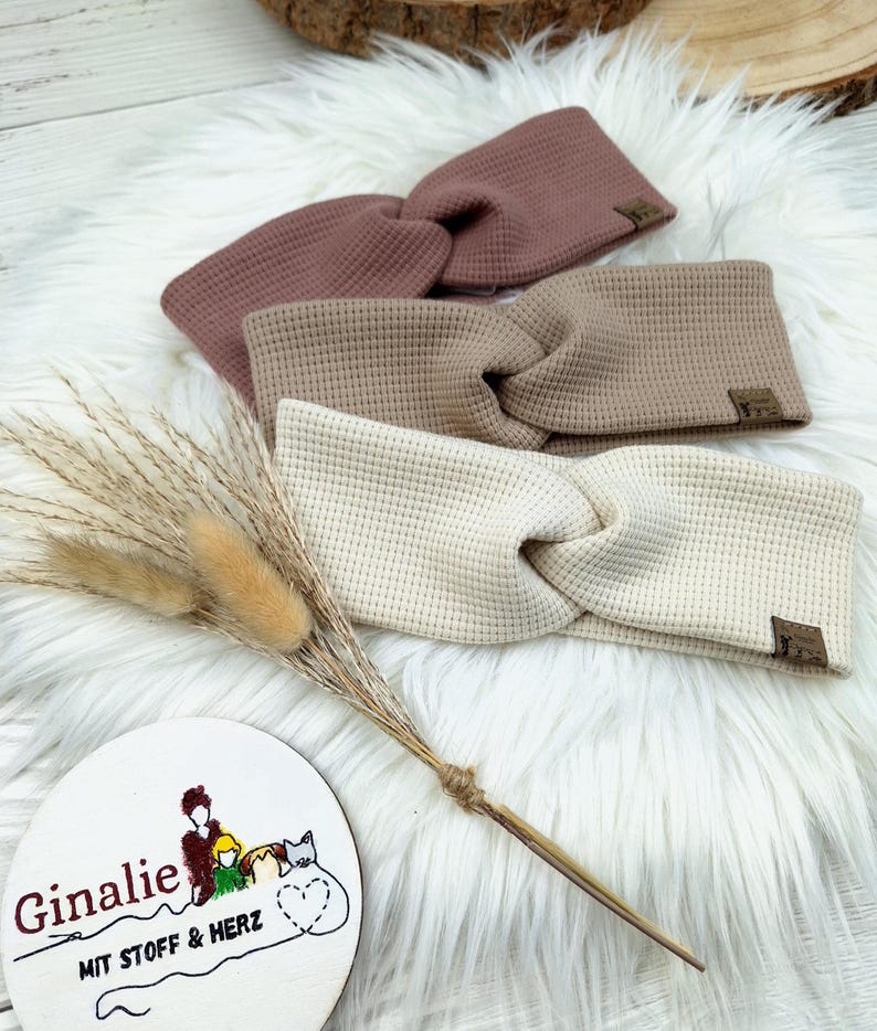 May include: Three waffle-knit headbands in shades of mauve, tan, and cream, each with a twisted knot design. Small brown tags are sewn on the headbands. The headbands are displayed on a white fluffy surface. A wooden sign with text and a drawing is in the foreground.