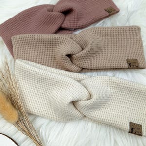 May include: Three waffle-knit headbands in shades of mauve, tan, and cream. Each headband features a twisted knot design and a small, rectangular label. The headbands are displayed on a fluffy, white surface.
