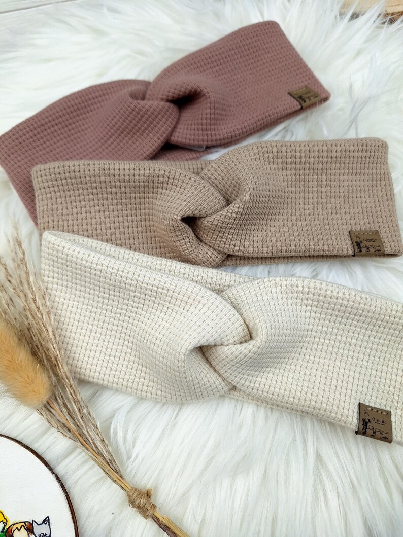 May include: Three waffle-knit headbands in shades of mauve, tan, and cream. Each headband features a twisted knot design and a small, rectangular label. The headbands are displayed on a fluffy, white surface.