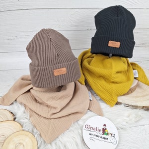 Op de afbeelding: Twee bruine en zwarte geribbelde gebreide mutsen met een bruin lederen label waarop "Ginalie mit Stoff & Herz" staat. De mutsen liggen op twee verschillende gekleurde sjaals. Eén sjaal is beige en de andere is geel.
