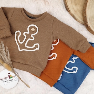 Op de afbeelding: Bruin wafelgebreid sweatshirt met een wit ankerontwerp en een houten knoopdetail. De top met lange mouwen maakt deel uit van een set met oranje en blauwe sweatshirts. De sweatshirts worden gepresenteerd op een witte, pluizige ondergrond.