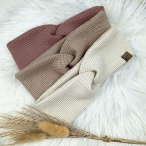 May include: Three waffle-knit headbands in shades of mauve, tan, and cream, arranged on a fluffy white surface. The headbands have a twisted knot design. A small brown label is visible on the cream headband. Dried grasses are in the foreground.